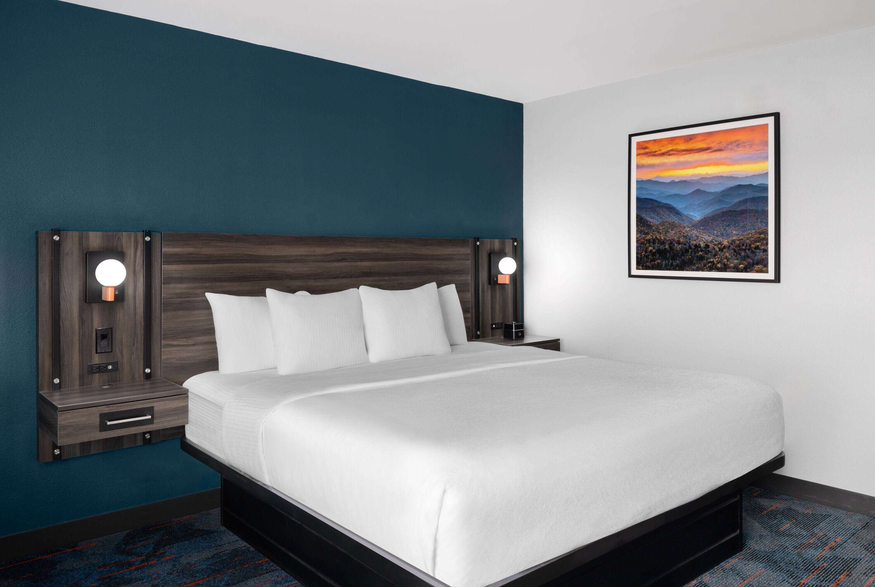 Habitación de hotel con una cama grande, dos mesitas de noche con lámparas, un cuadro enmarcado de un paisaje en la pared y una pared de acento azul oscuro.