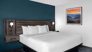 A hotel room with a large bed, two bedside tables with lamps, a framed landscape painting on the wall, and a dark blue accent wall.