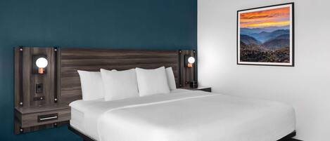 Habitación de hotel con una cama grande, dos mesitas de noche con lámparas, un cuadro enmarcado de un paisaje en la pared y una pared de acento azul oscuro.