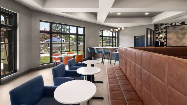 Un área de estar interior moderna con mesas y sillas, un banco acolchado y amplios ventanales que ofrecen vistas al exterior.