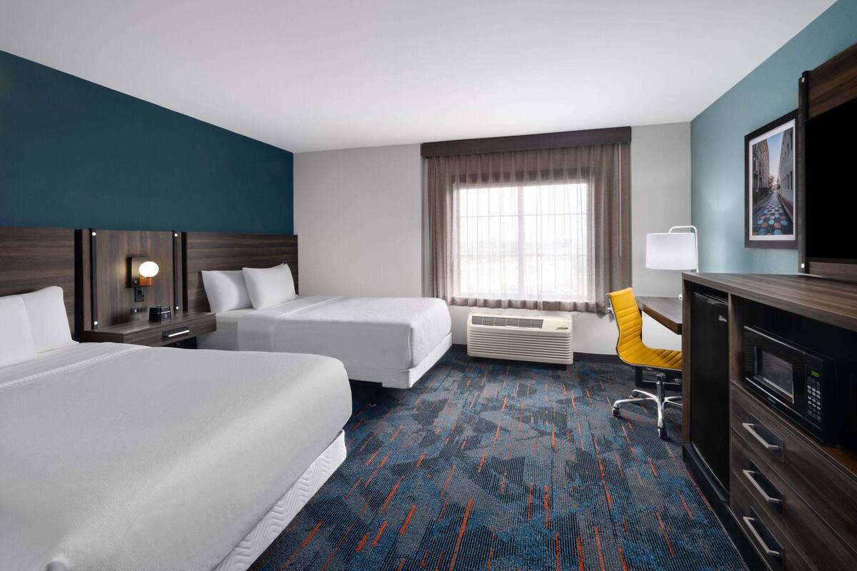 A hotel room with two beds, a desk with a chair, a TV, an air conditioning unit, and a large window with curtains.