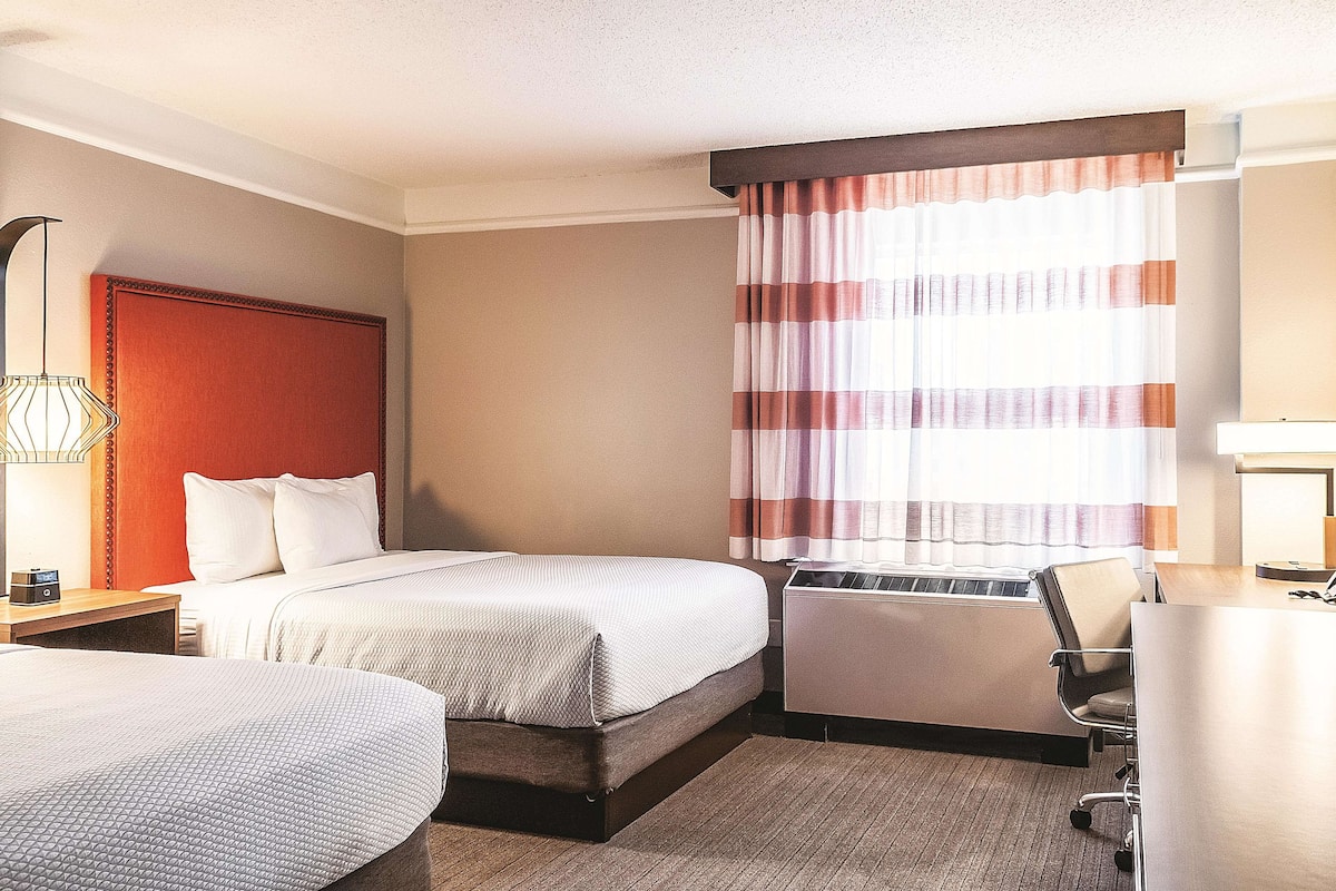 A hotel room with two beds, a desk, a chair, a heater, and a window with curtains.