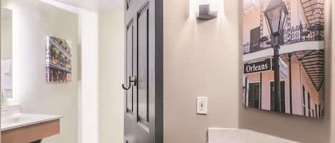 A hotel bathroom with a white sink, a door with a handle, a wall-mounted light, and a poster of a cityscape.