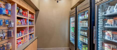 A narrow hallway with wooden shelves stocked with snacks and beverages, a chandelier, and two refrigerators filled with drinks.