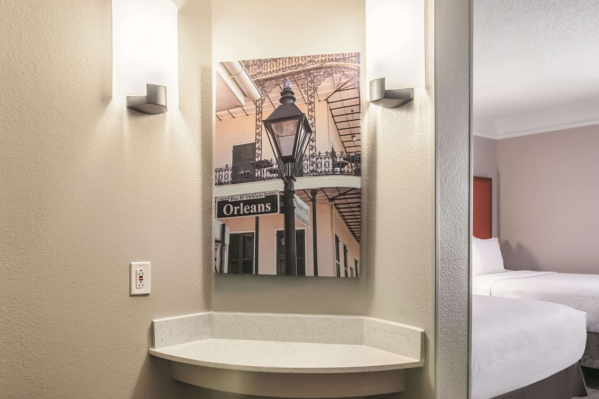 A hotel bathroom with a corner sink, two wall-mounted lights, and a mirror with a cityscape print.