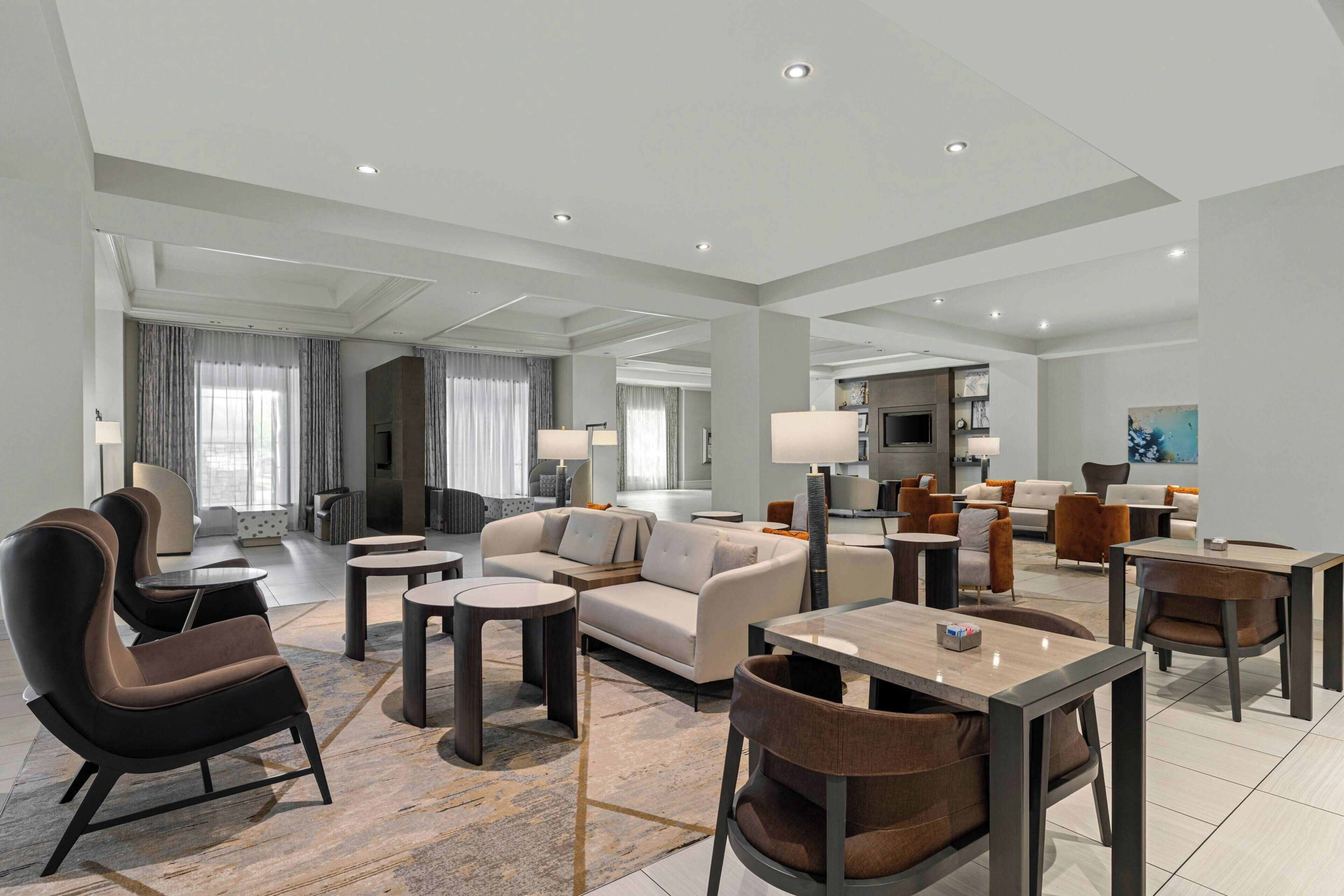 A spacious hotel lobby with multiple seating areas, including sofas and chairs, a dining table, and a kitchen area visible in the background.