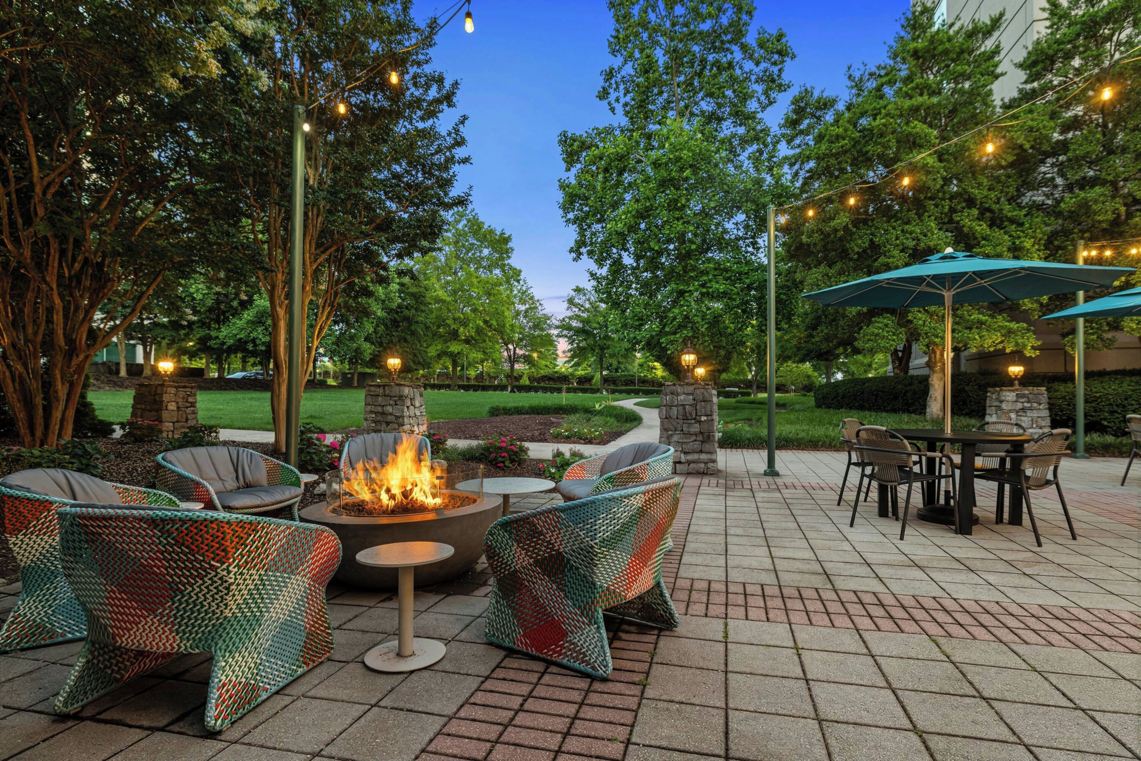 An outdoor patio with wicker furniture, a central fire pit, stone pillars, and string lights.