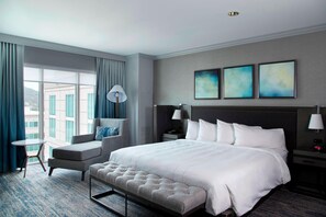 A modern hotel room with a large bed, a seating area with a chair and a small table, and a large window offering a city view.