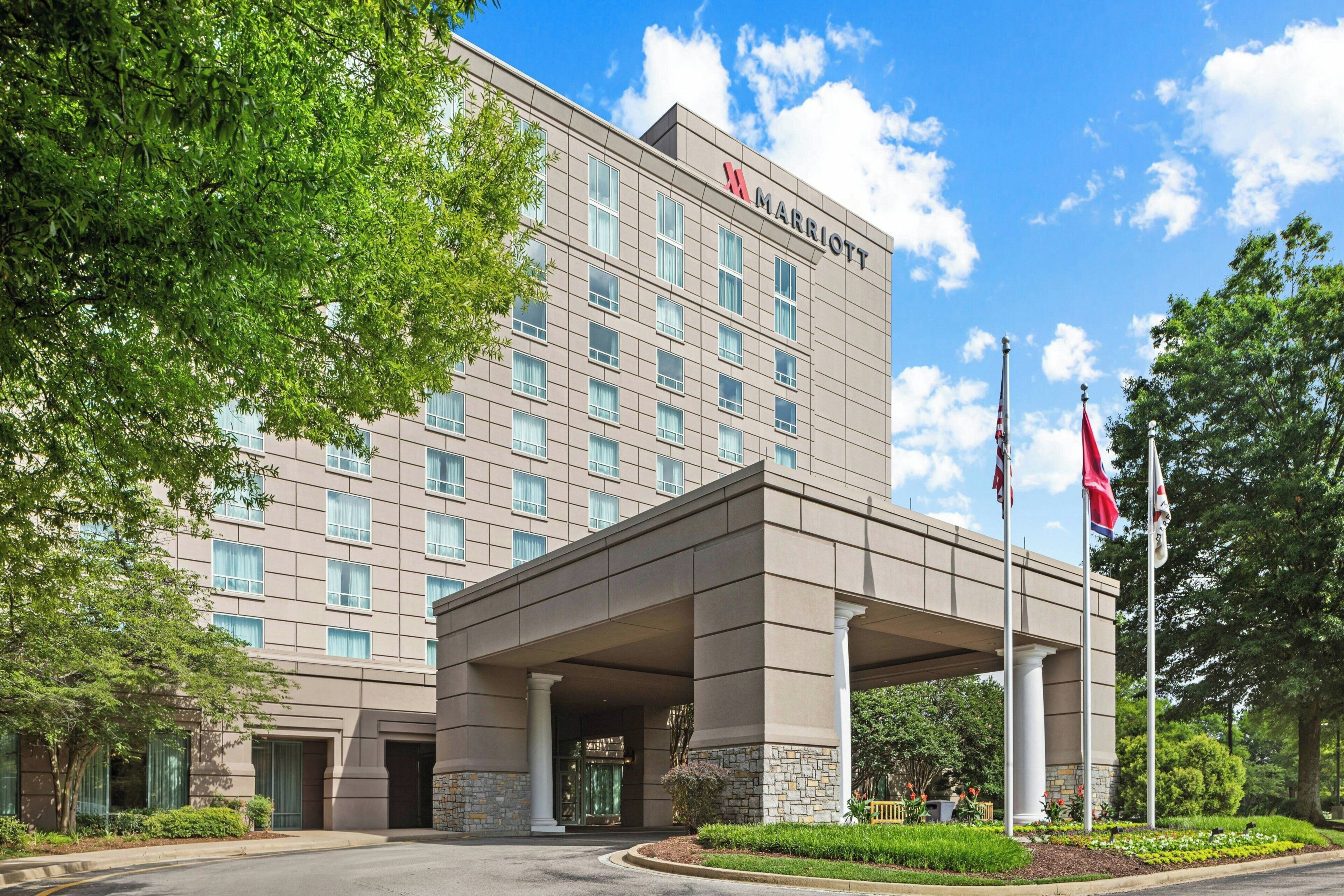 Un hotel de varios pisos con una entrada destacada, asta de banderas y un área ajardinada con árboles y arbustos.