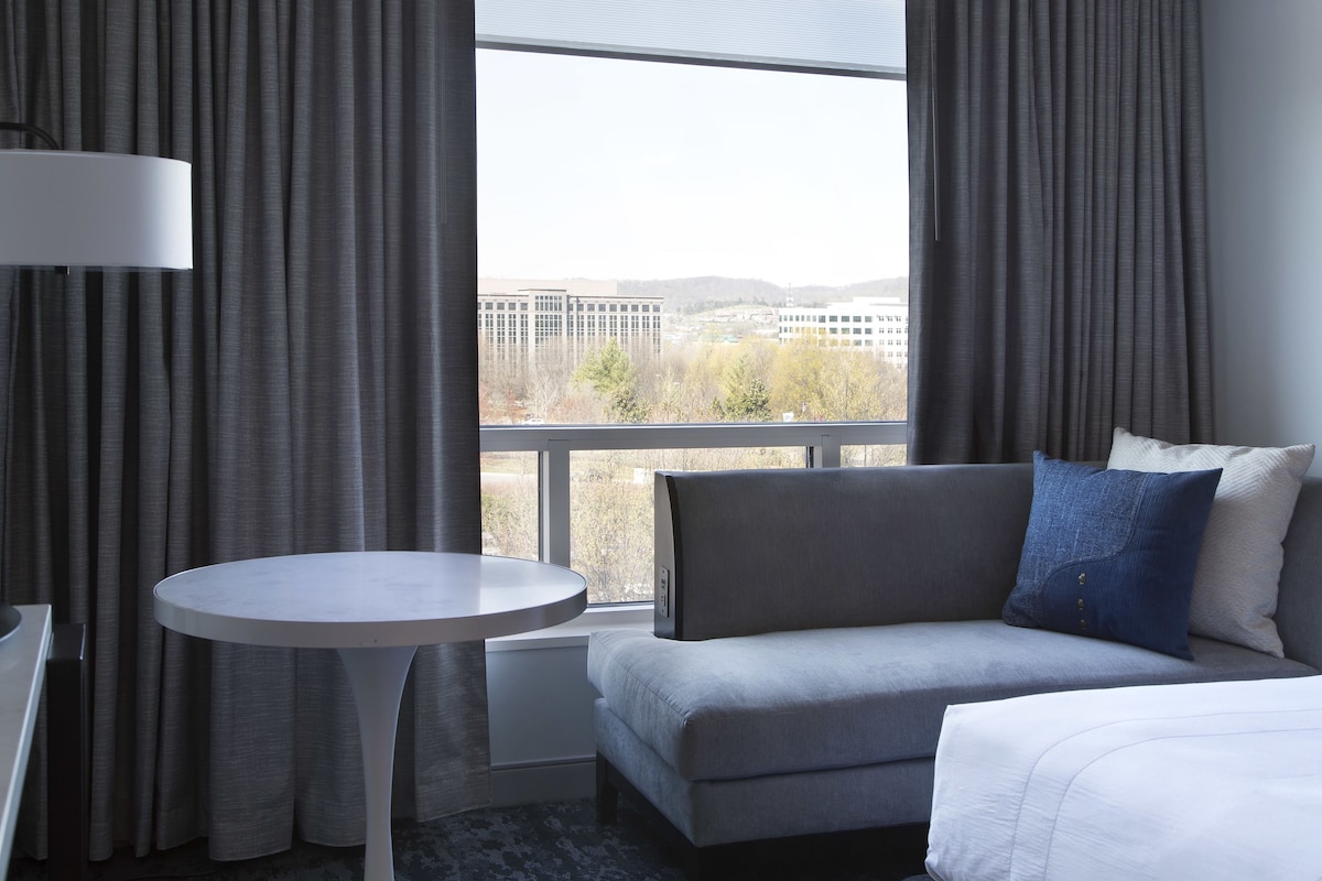 A modern hotel room with a grey sofa, a small round table, and a large window offering a city view.