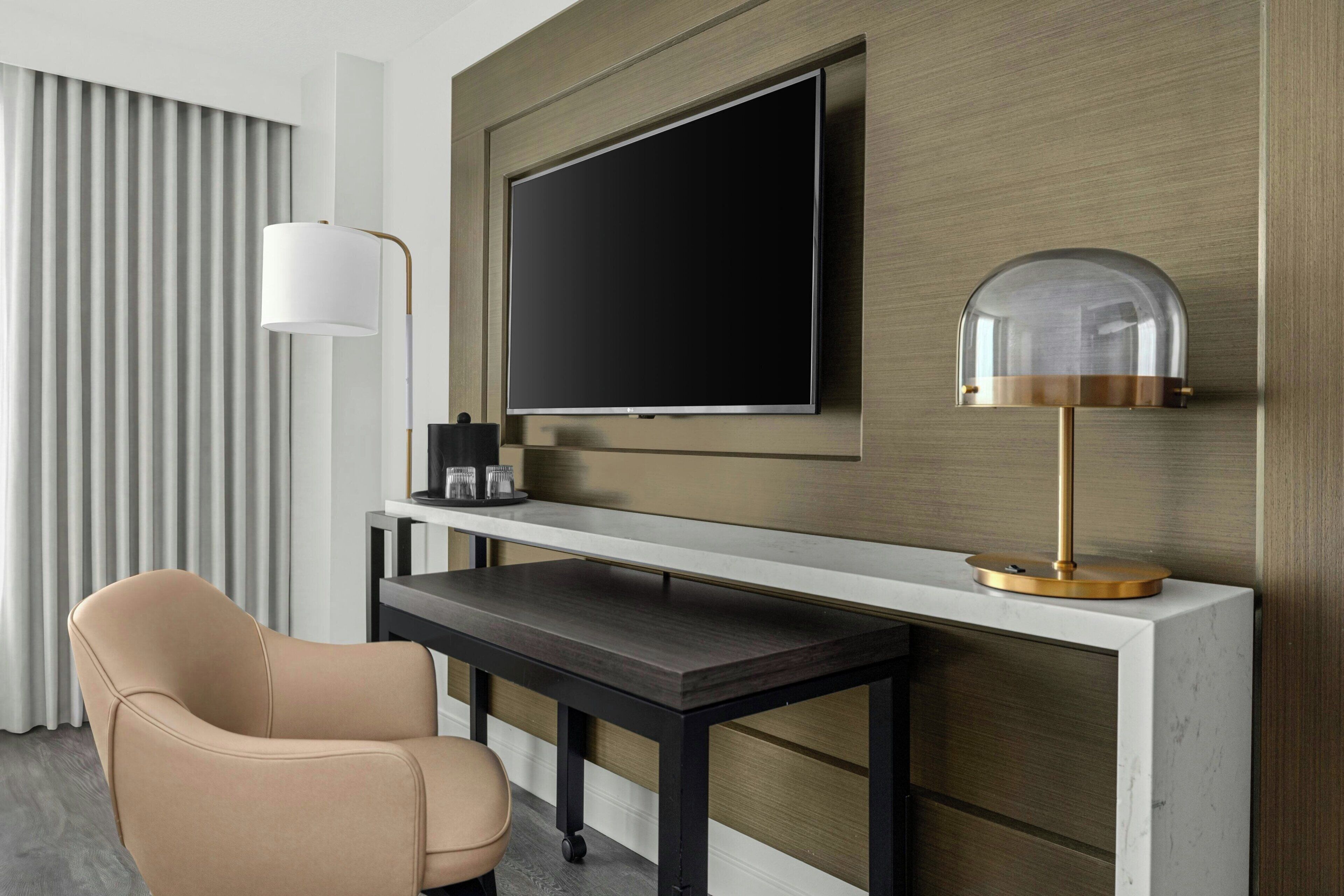 A modern hotel room with a flat-screen TV mounted on a wooden panel, a desk with a lamp, and a leather chair.