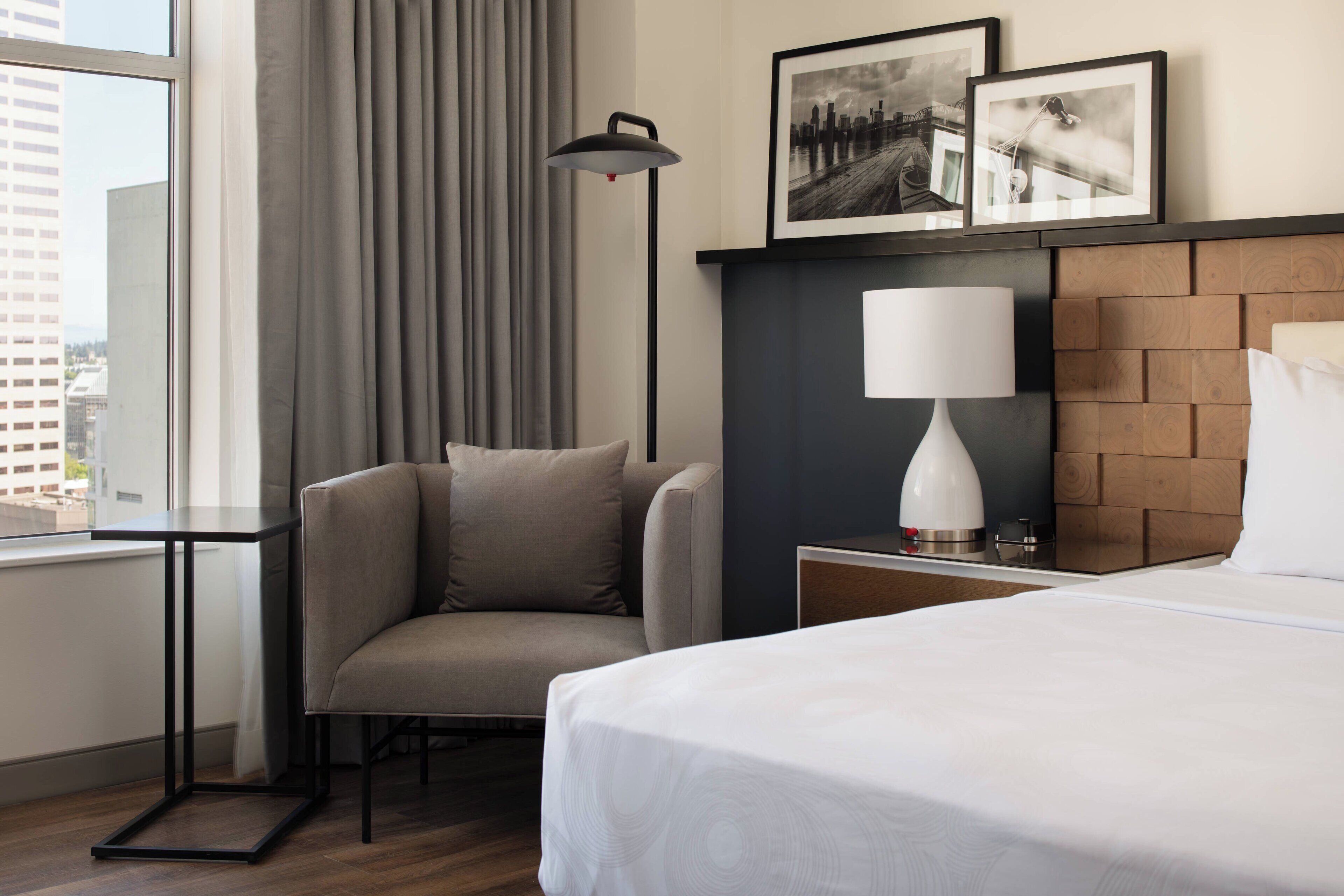 A hotel room with a bed, a chair, a desk, a lamp, and two framed pictures on the wall.