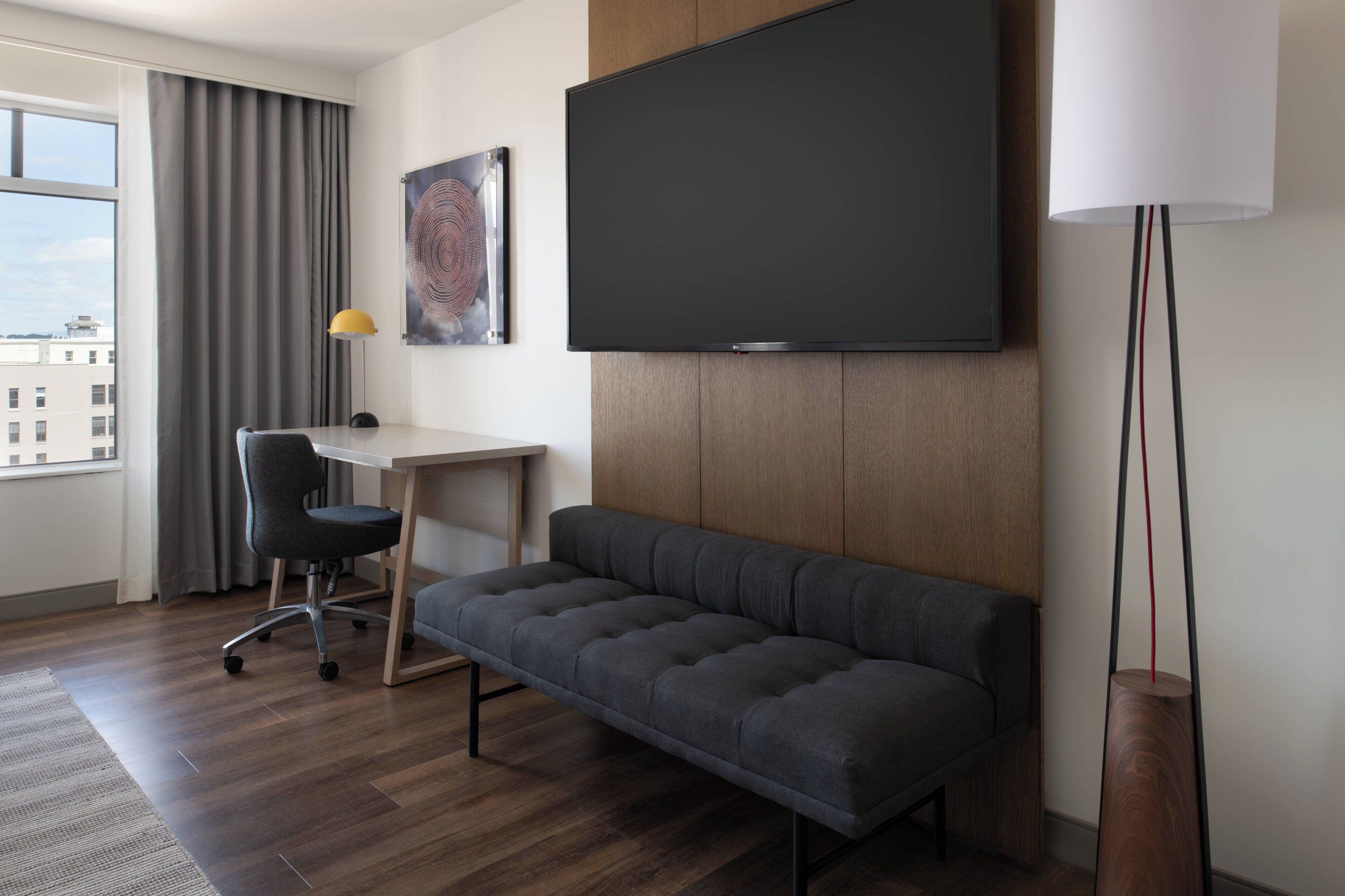 Une chambre d’hôtel moderne dotée d’un grand téléviseur à écran plat, d’un canapé gris, d’un bureau avec une chaise et d’une lampe sur pied.