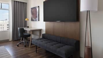 Une chambre d’hôtel moderne dotée d’un grand téléviseur à écran plat, d’un canapé gris, d’un bureau avec une chaise et d’une lampe sur pied.