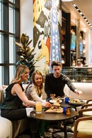 Un gruppo di persone che cena a un tavolo in un ambiente interno moderno, con ampie finestre e un affresco sulla parete.
