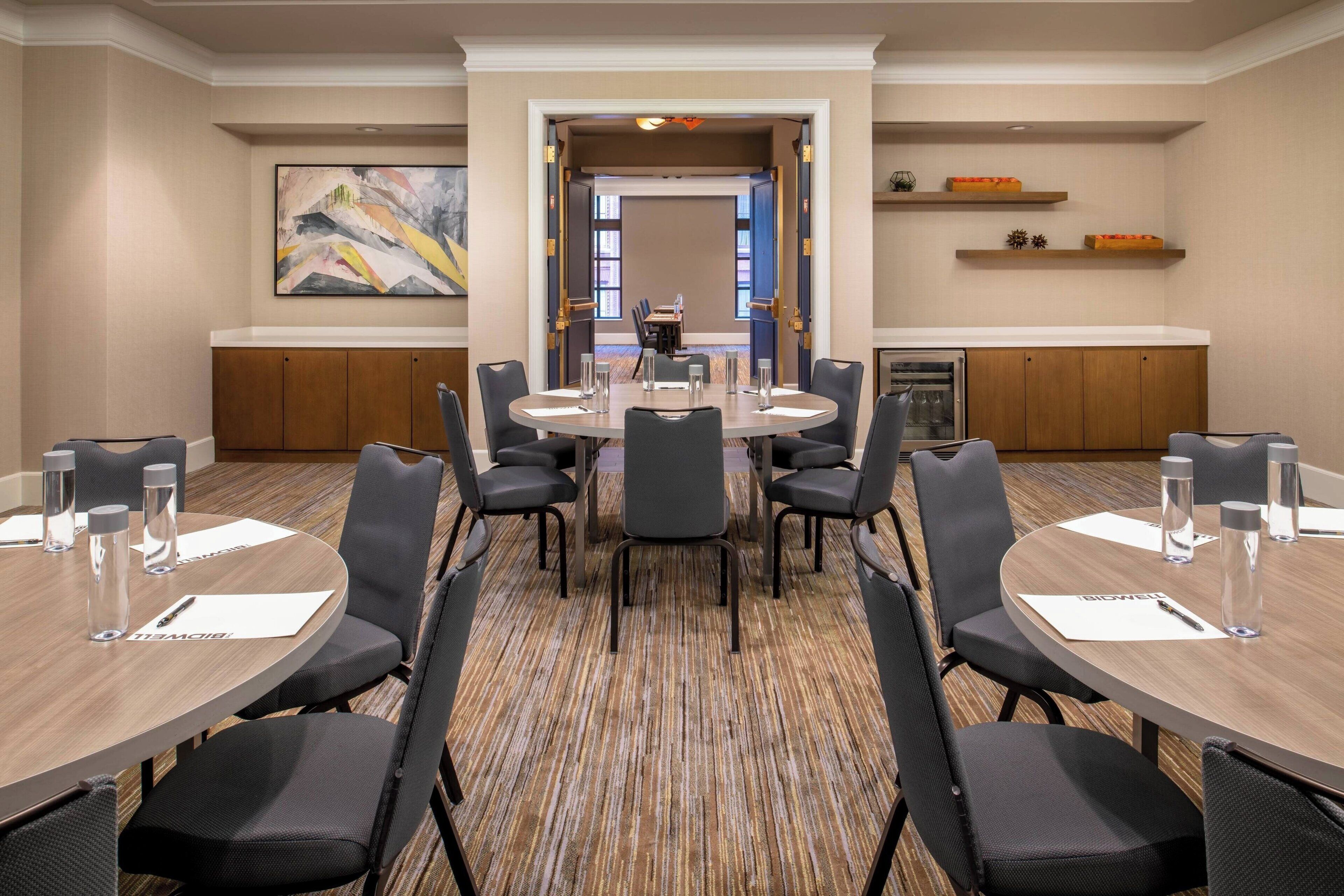A conference room with a round table, black chairs, and water bottles. There is a painting on the wall and a kitchen area in the background.