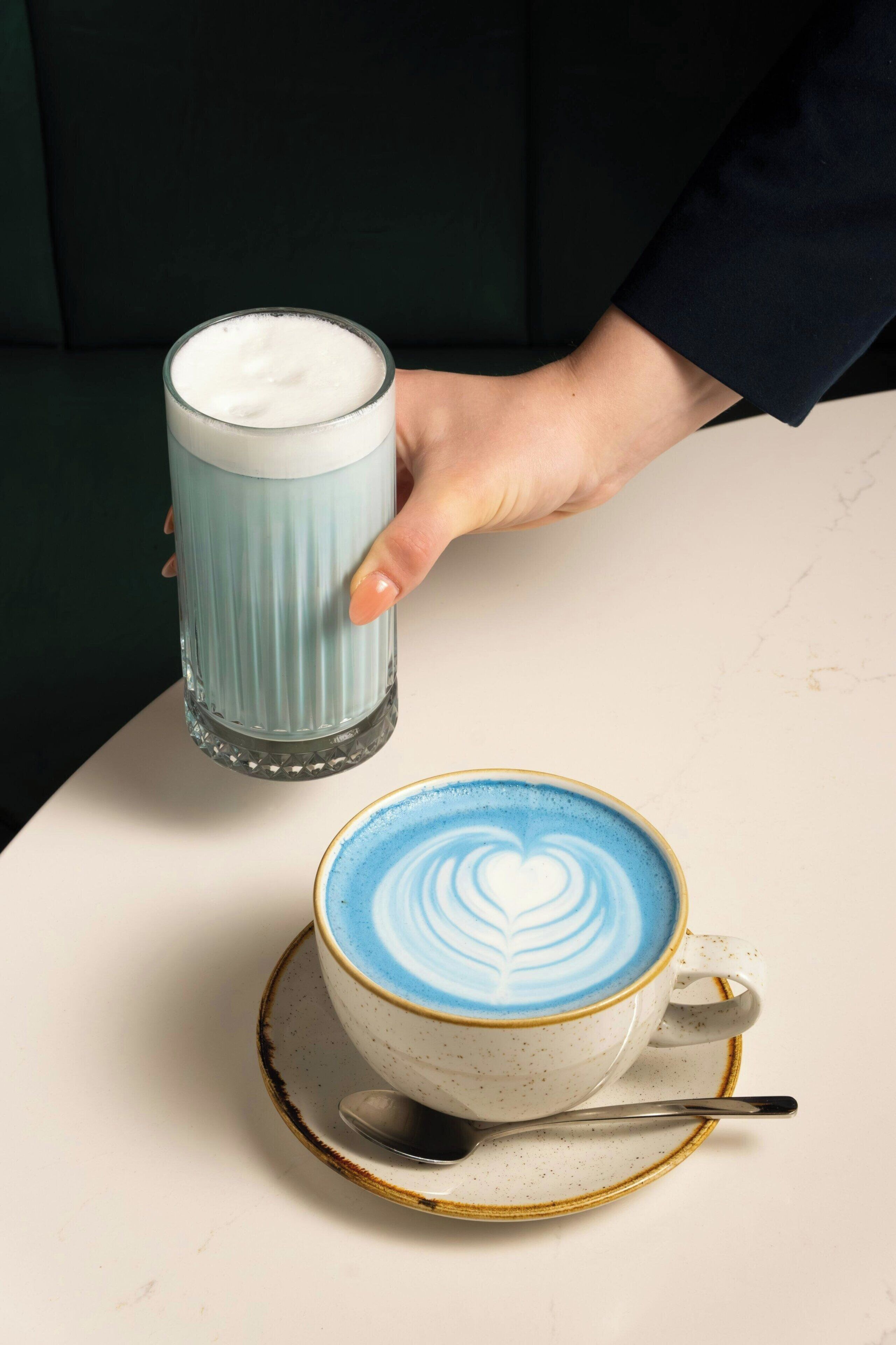 Uma mão segurando um copo de leite, uma xícara de café com arte de latte e uma colher em um pires.