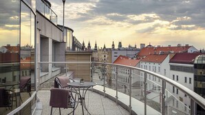 Un balcón con barandilla de vidrio, una mesa y dos sillas, con vistas a un paisaje urbano que incluye edificios históricos y un cielo nublado.
