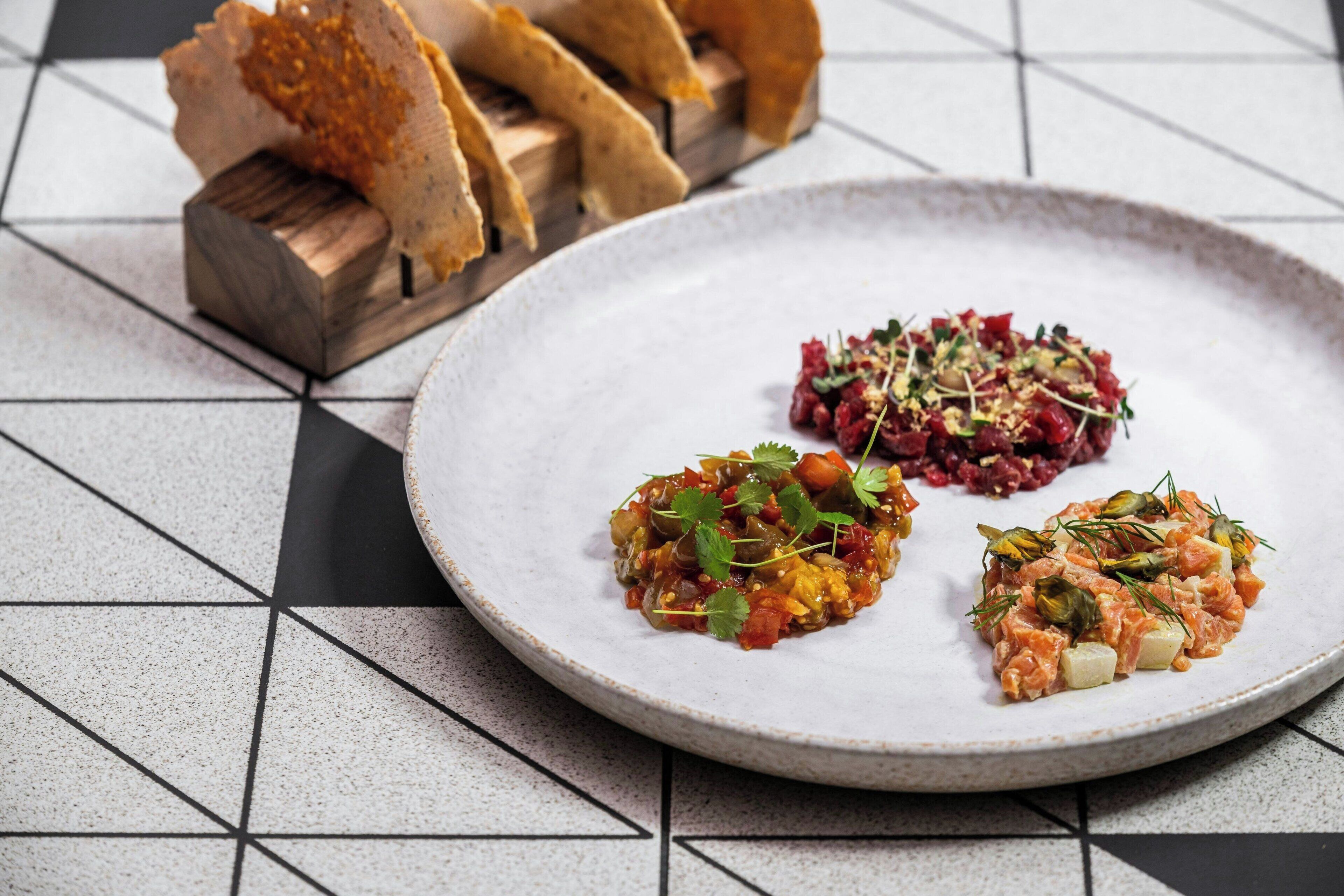 A plate with three different dishes, a wooden board with breadsticks, and a tiled floor.