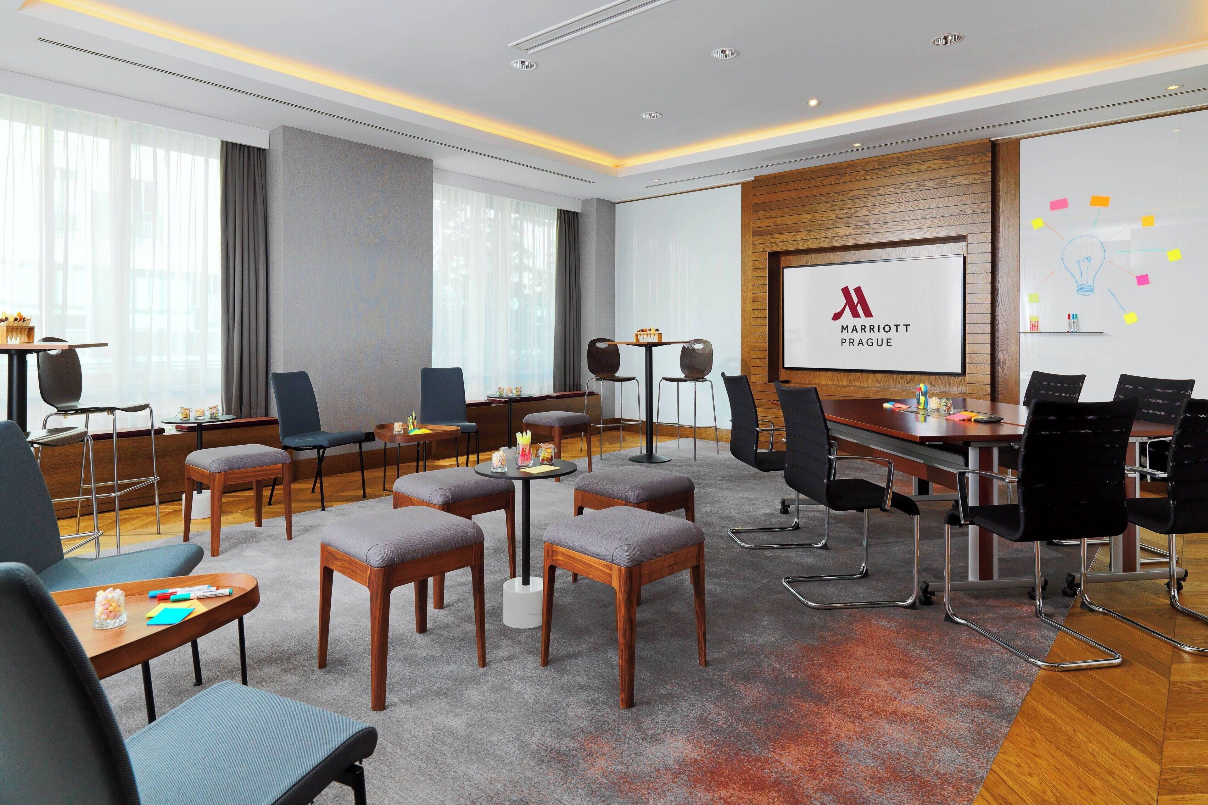 Et moderne mødelokale med et stort skærmbillede af Marriott Prague logoet, flere forskellige siddepladser og en trævæg.