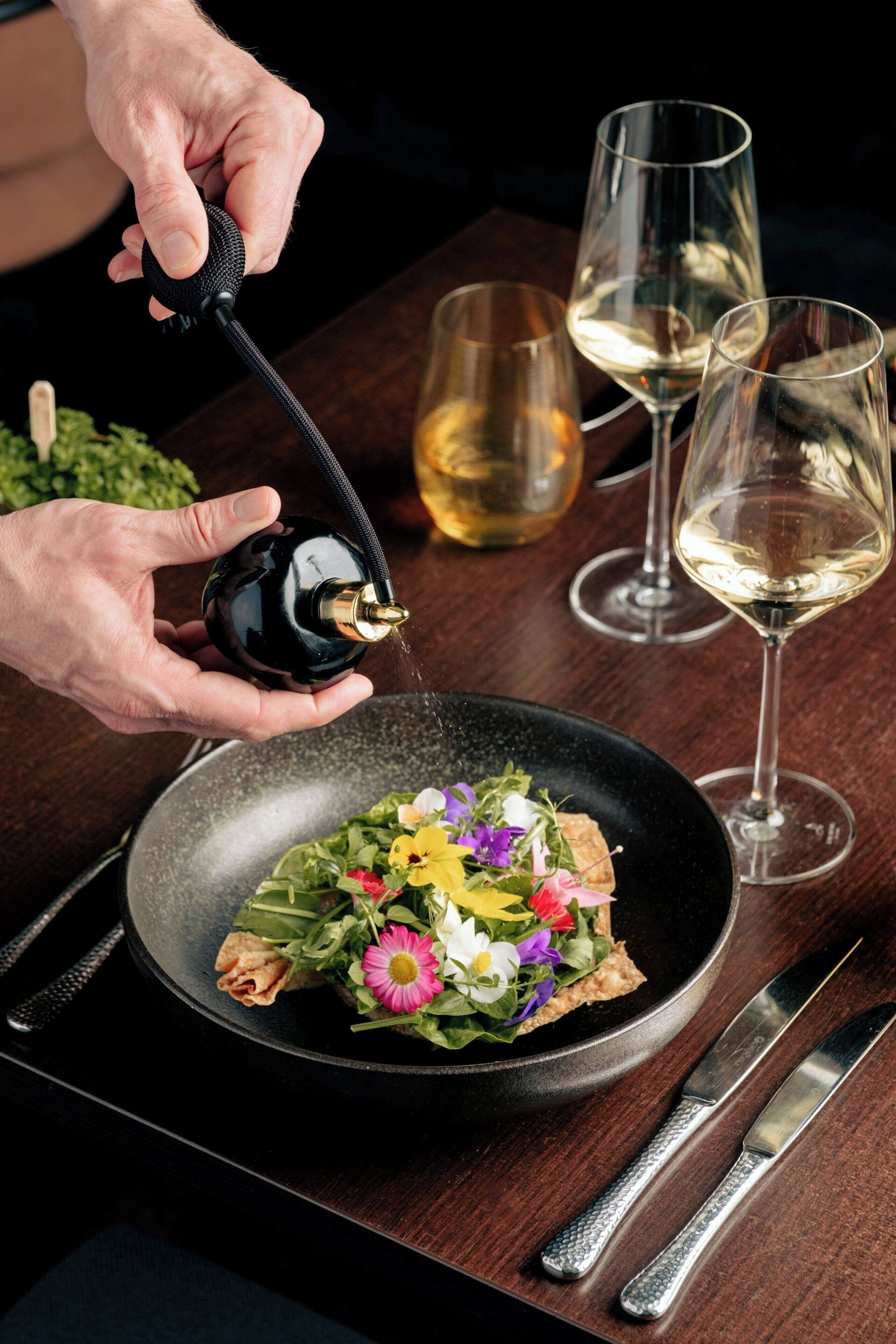 Una persona sirve agua de una botella en un bol de ensalada con flores. Hay dos copas de vino blanco sobre la mesa.