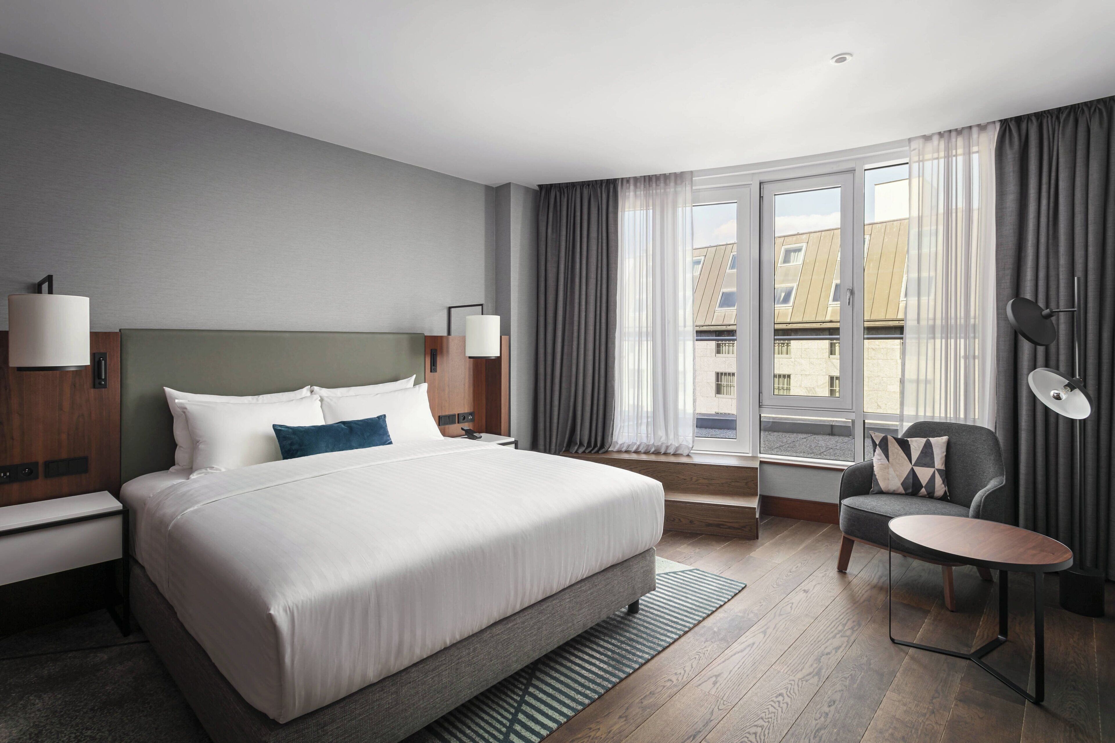 Habitación de hotel con una cama grande, un escritorio, una silla y vistas a la ciudad por la ventana.