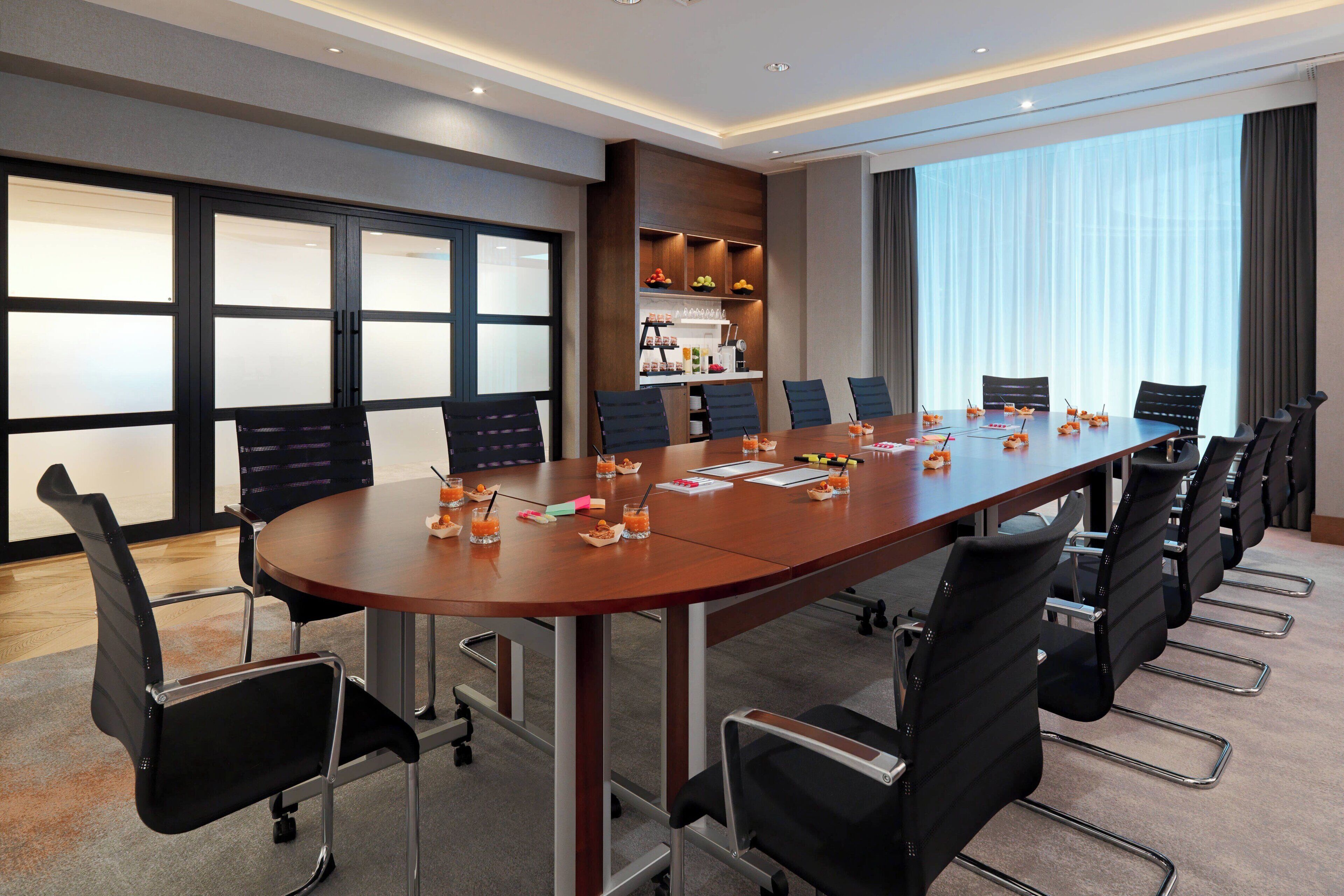 A conference room with a long table, multiple chairs, and a kitchenette area with shelves.
