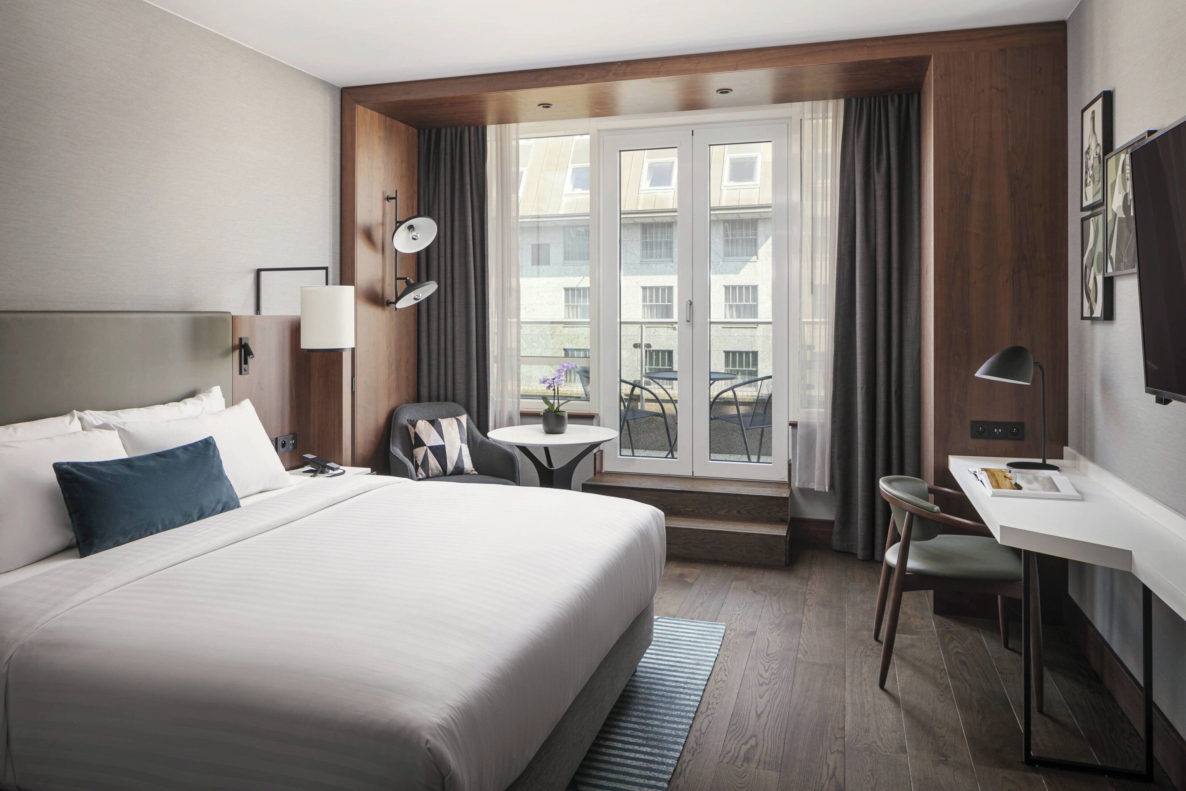 A hotel room with a large bed, a desk, a chair, a small table with a plant, and a balcony with a view of buildings.
