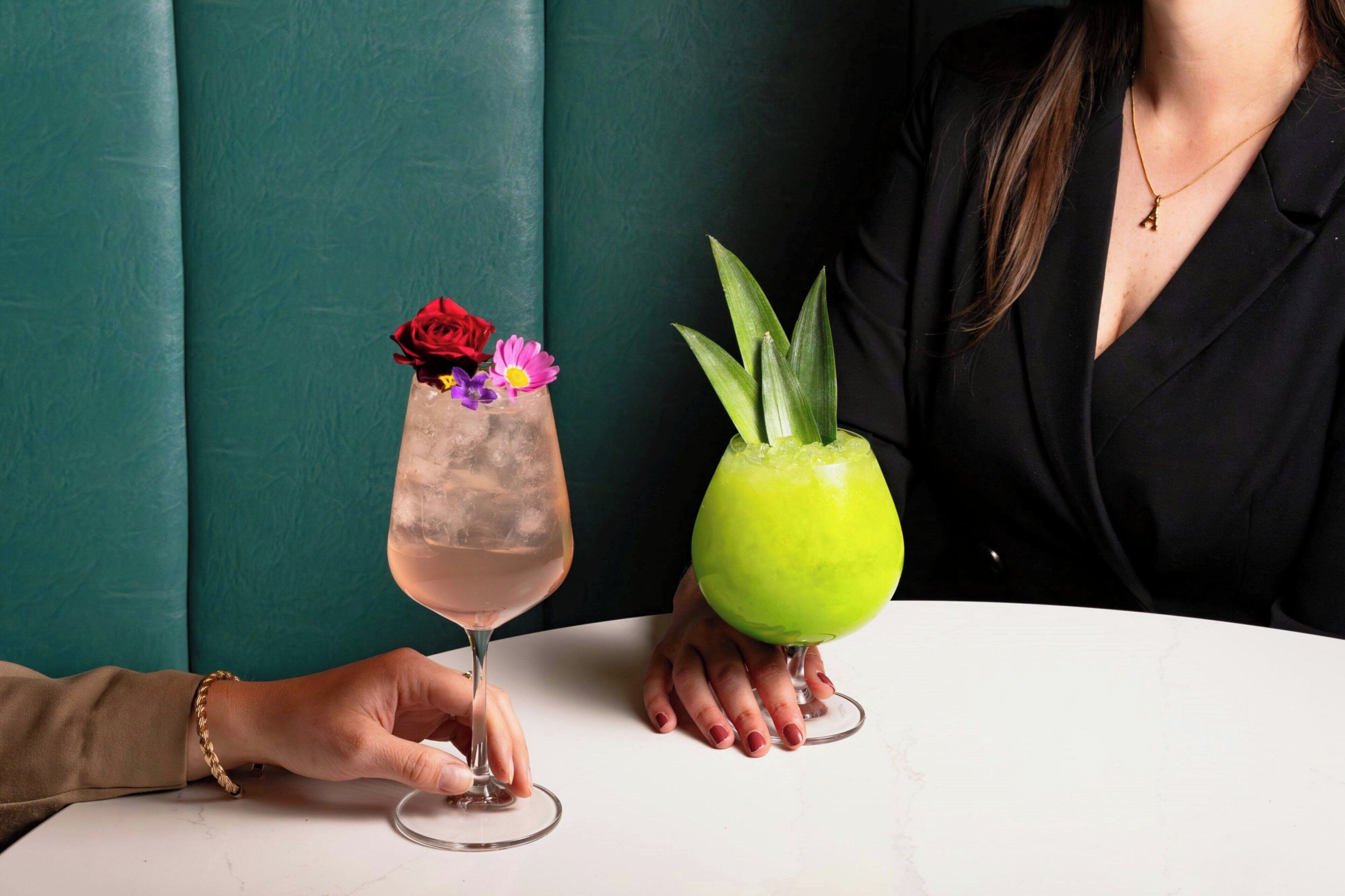 Una persona sosteniendo un vaso con una rosa y flores, y otro vaso con una bebida verde y una planta.