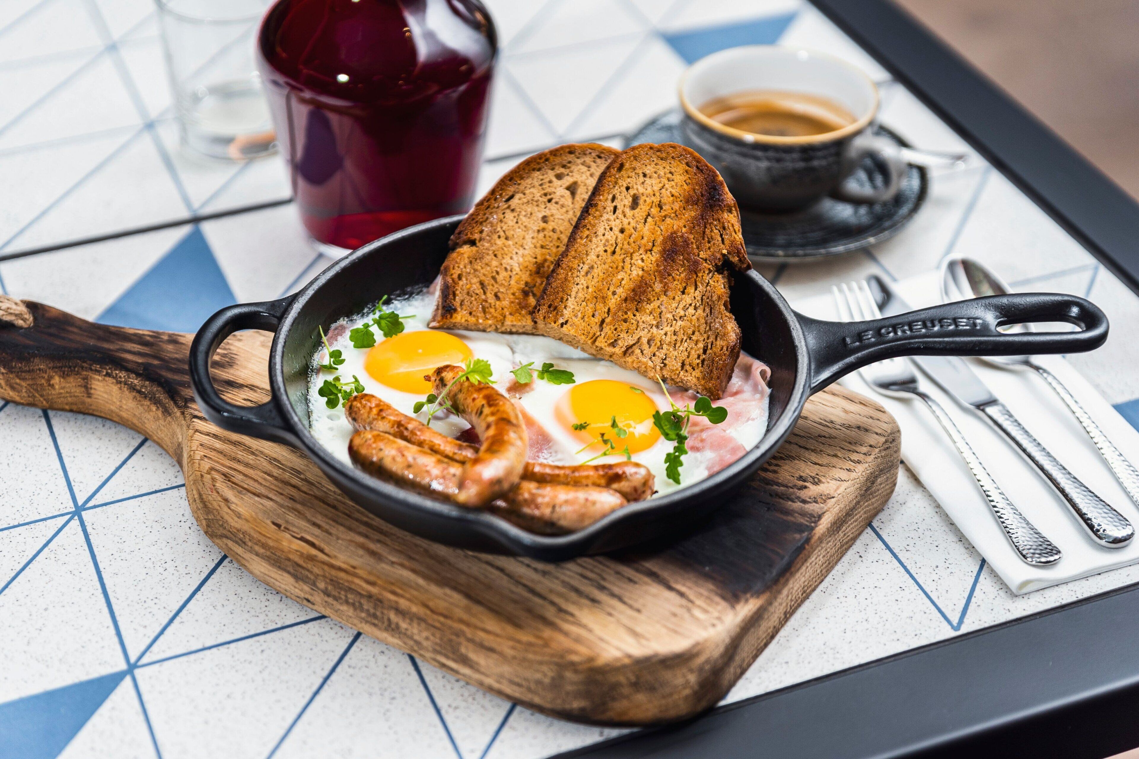 A breakfast setting with a skillet containing eggs, sausages, and toast on a wooden board.
