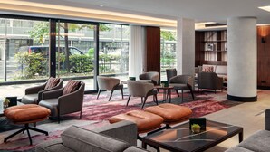Un moderno vestíbulo de hotel con sofás de cuero, sillones y una mesa de centro. Amplias ventanas ofrecen vistas a la calle y vehículos estacionados.