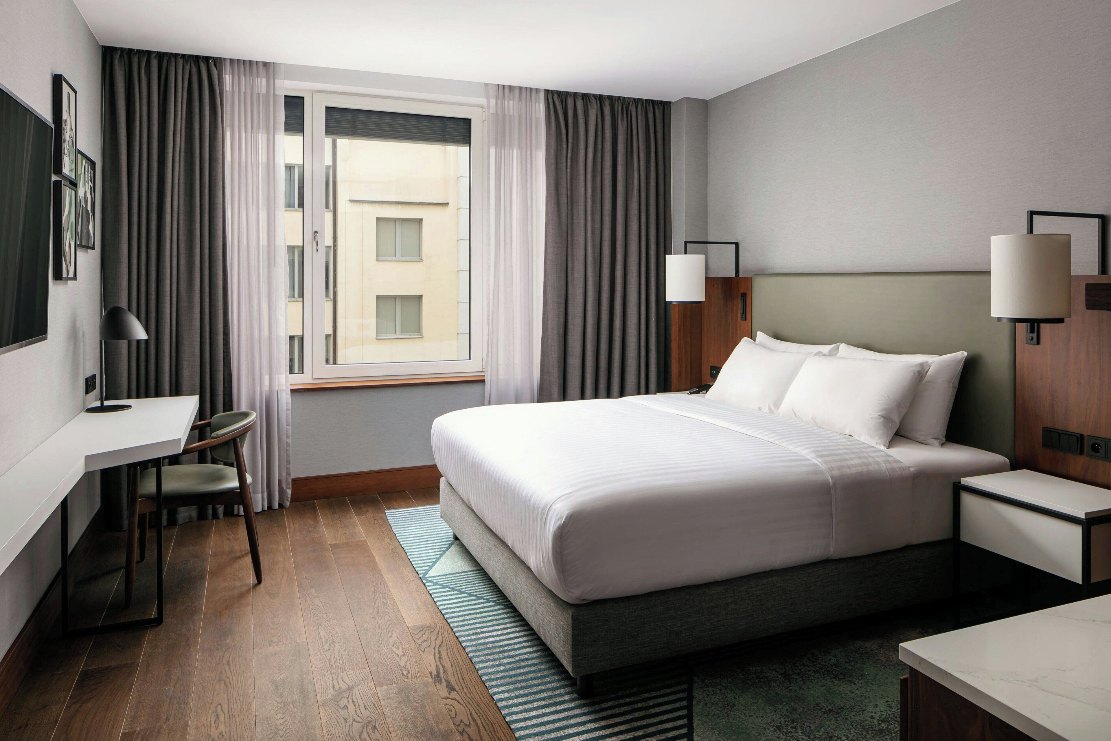 A hotel room with a large bed, a desk, a chair, two bedside tables, and a window with curtains.