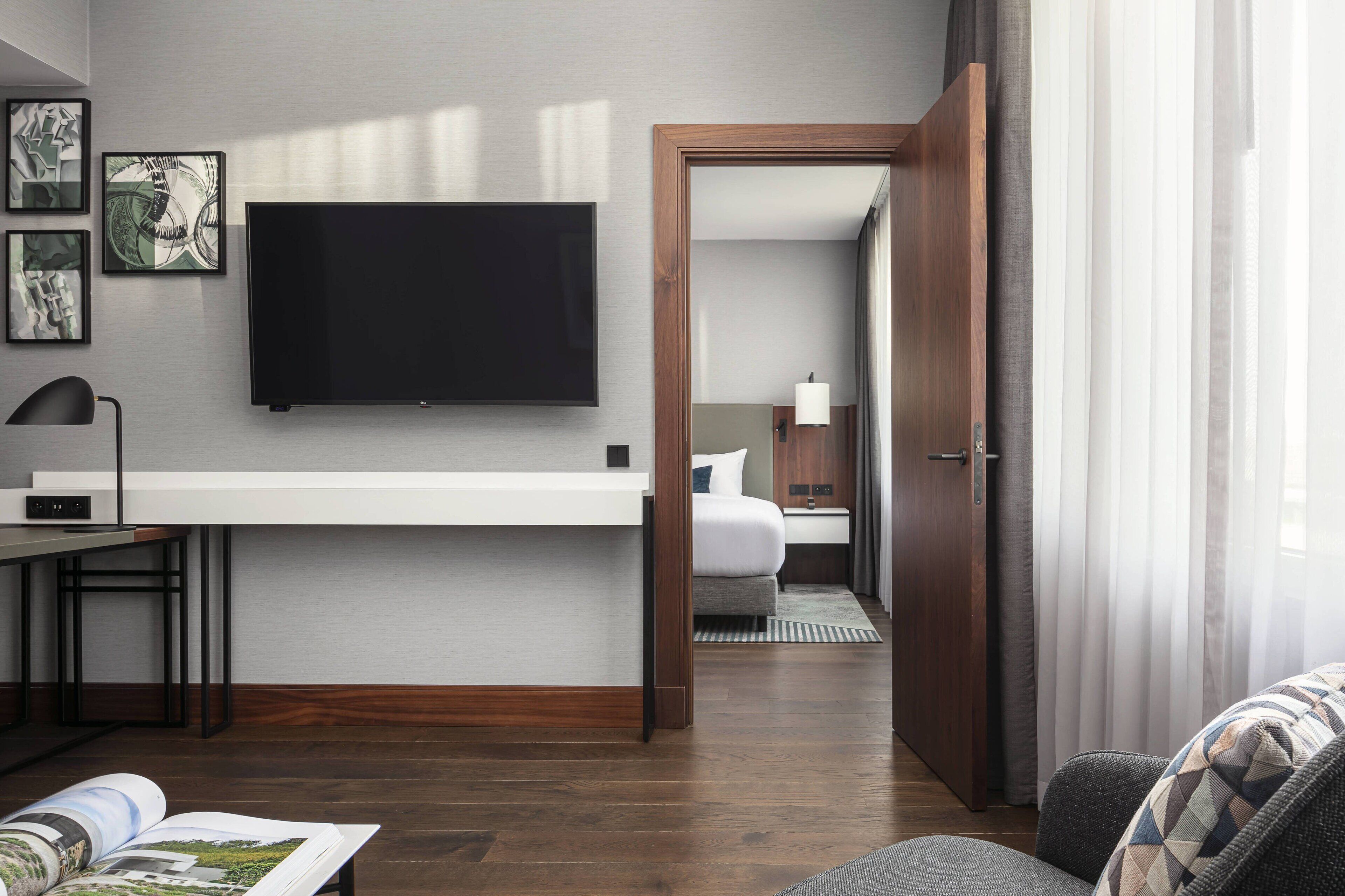 Quarto de hotel moderno com televisão de tela plana na parede, uma escrivaninha de madeira com luminária, uma cama com mesa de cabeceira e vista através de uma porta aberta.