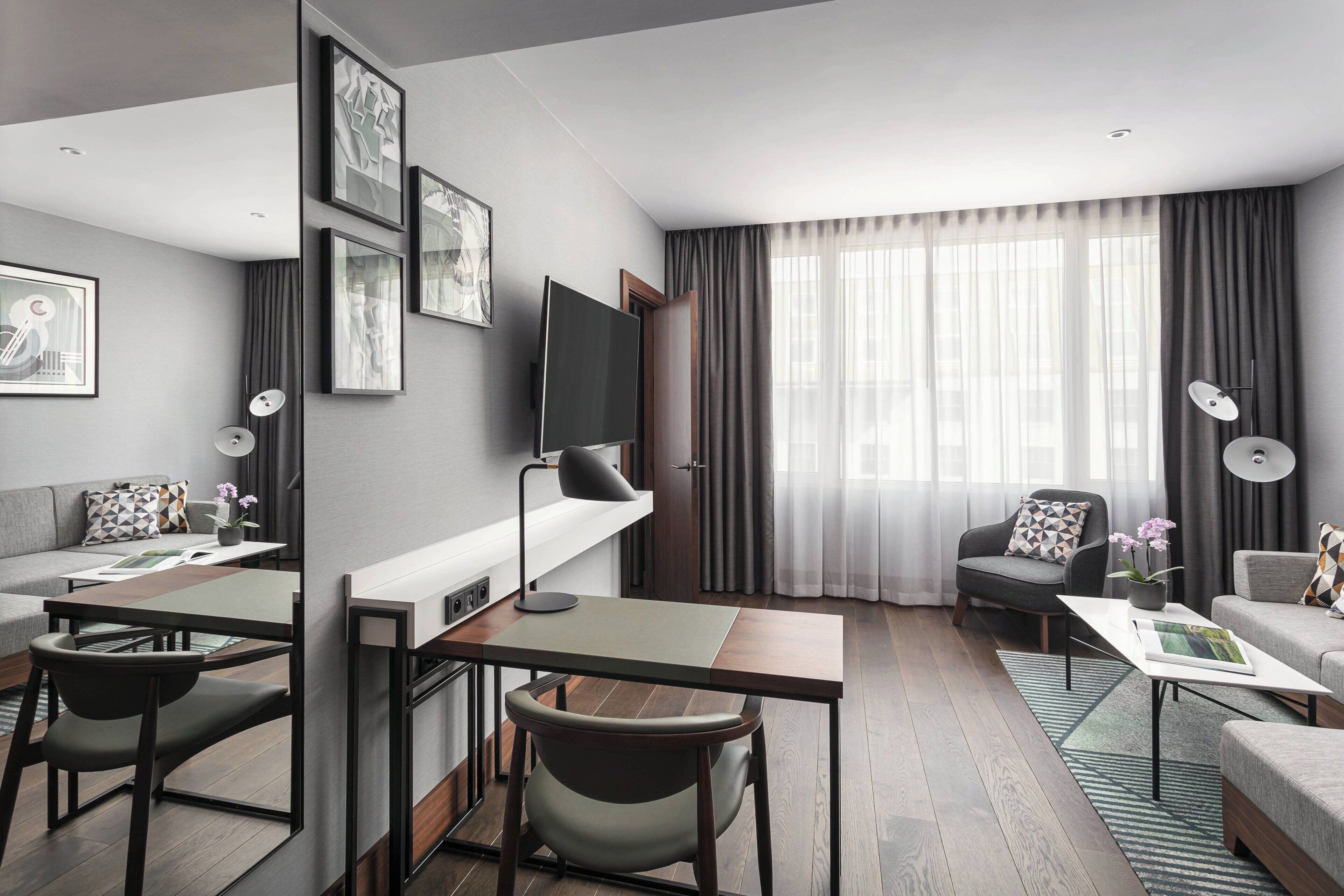 Une chambre d’hôtel moderne, équipée d’un canapé, d’un bureau avec une chaise, d’une télévision et d’une grande fenêtre avec des rideaux.