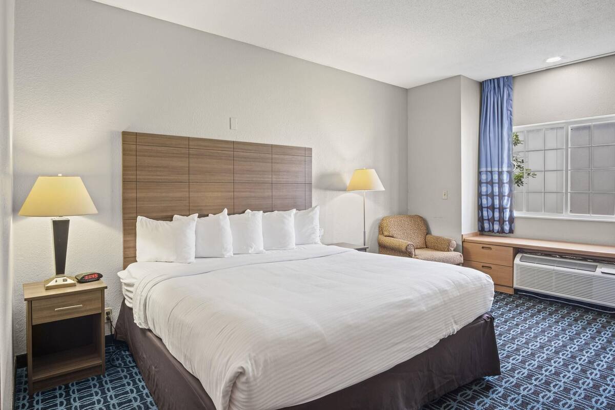A hotel room with a large bed, two bedside lamps, a desk with a chair, a window with curtains, and a radiator.