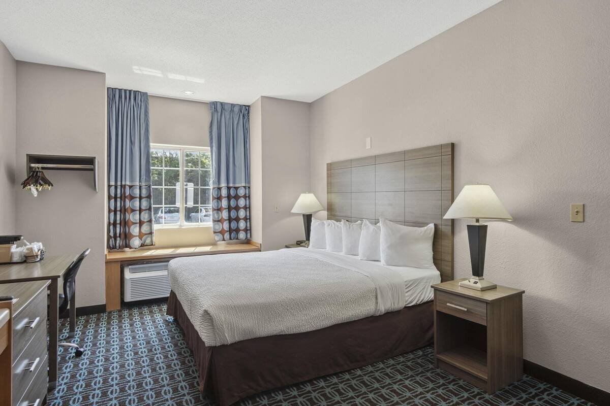 A hotel room with a bed, two bedside tables with lamps, a desk with a chair, a window with curtains, and a wall-mounted heater.