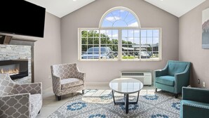 A living room with a fireplace, patterned chairs, a coffee table, and a large window with a view of parked cars and greenery.