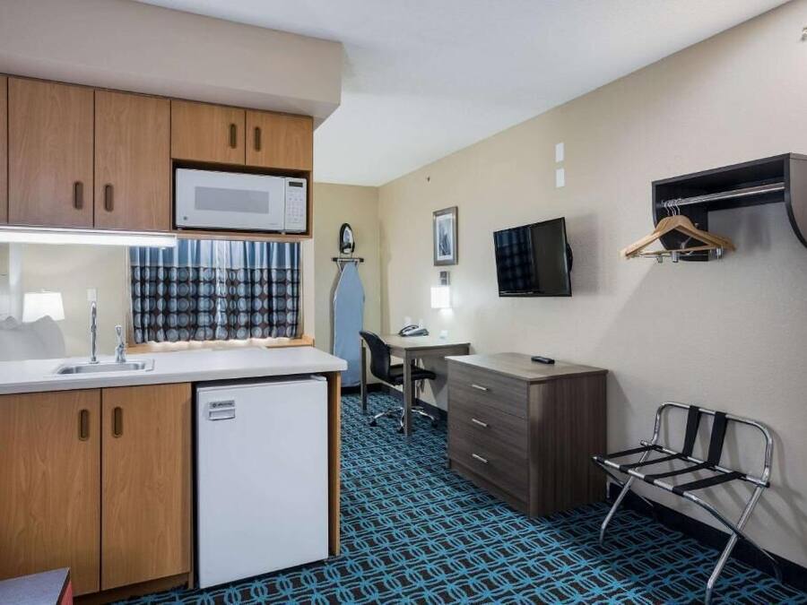 Quarto de hotel compacto com cozinha compacta, escrivaninha, cadeira, televisão na parede e um pequeno espaço para sentar.