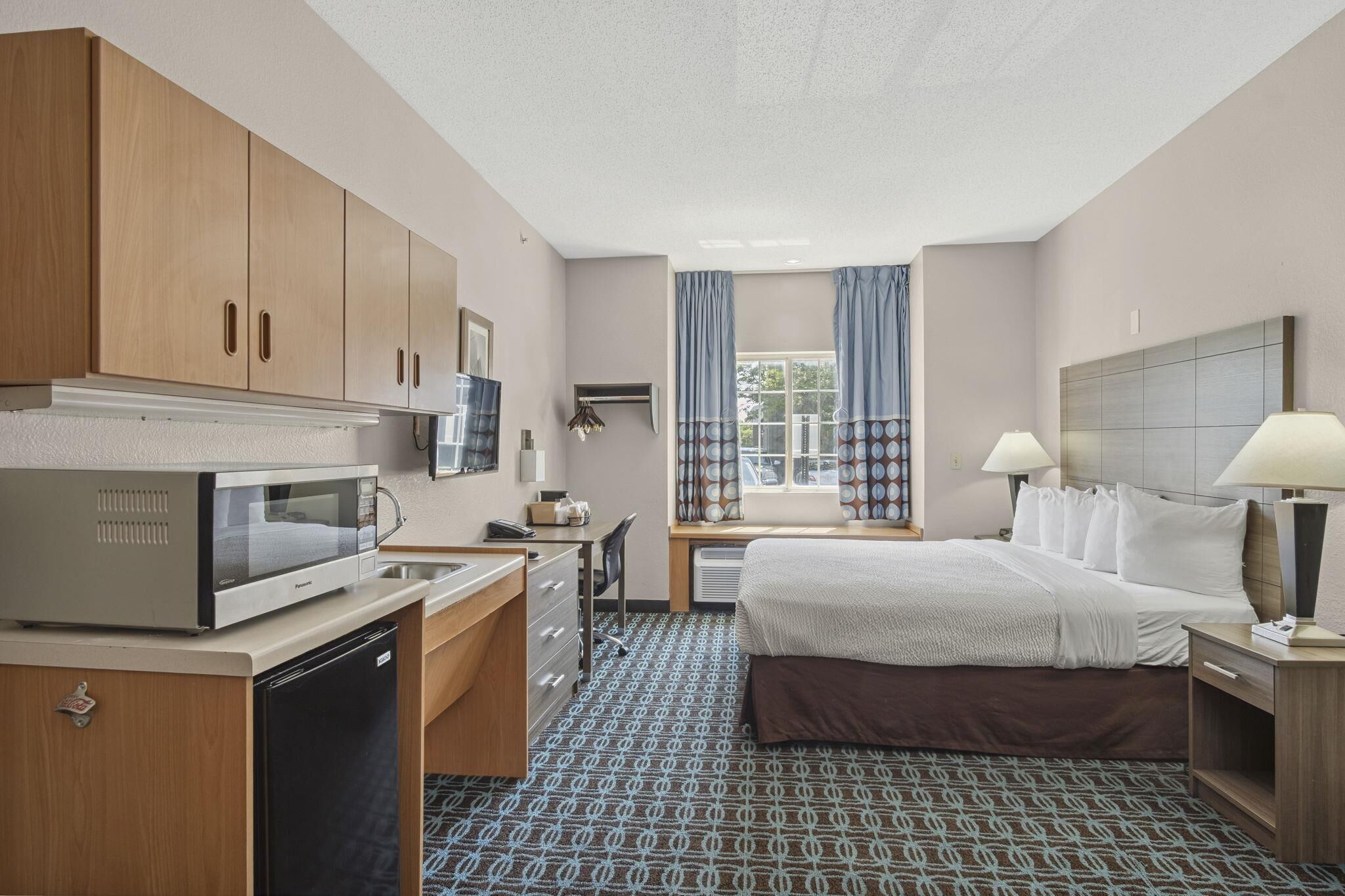 A hotel room with a kitchenette, a bed, a desk, and a window with curtains.