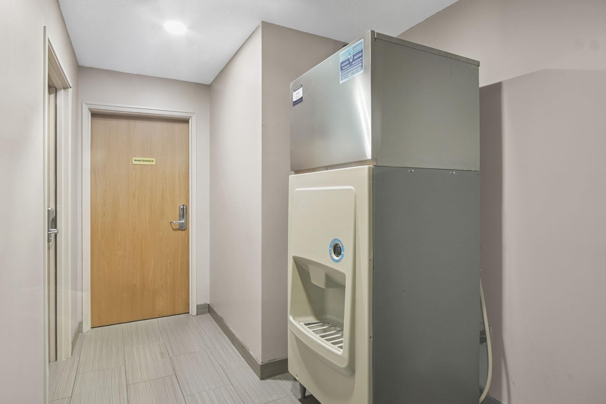 Un pasillo con una puerta de madera que indica 'Mantenimiento', un refrigerador y un dispensador de agua.