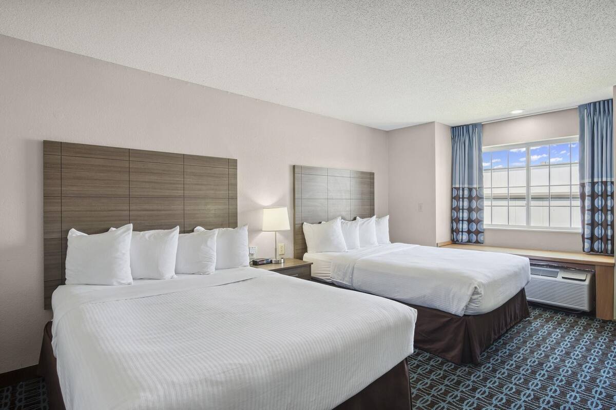 A hotel room with two beds, a large window with curtains, and a headboard with a built-in heater.