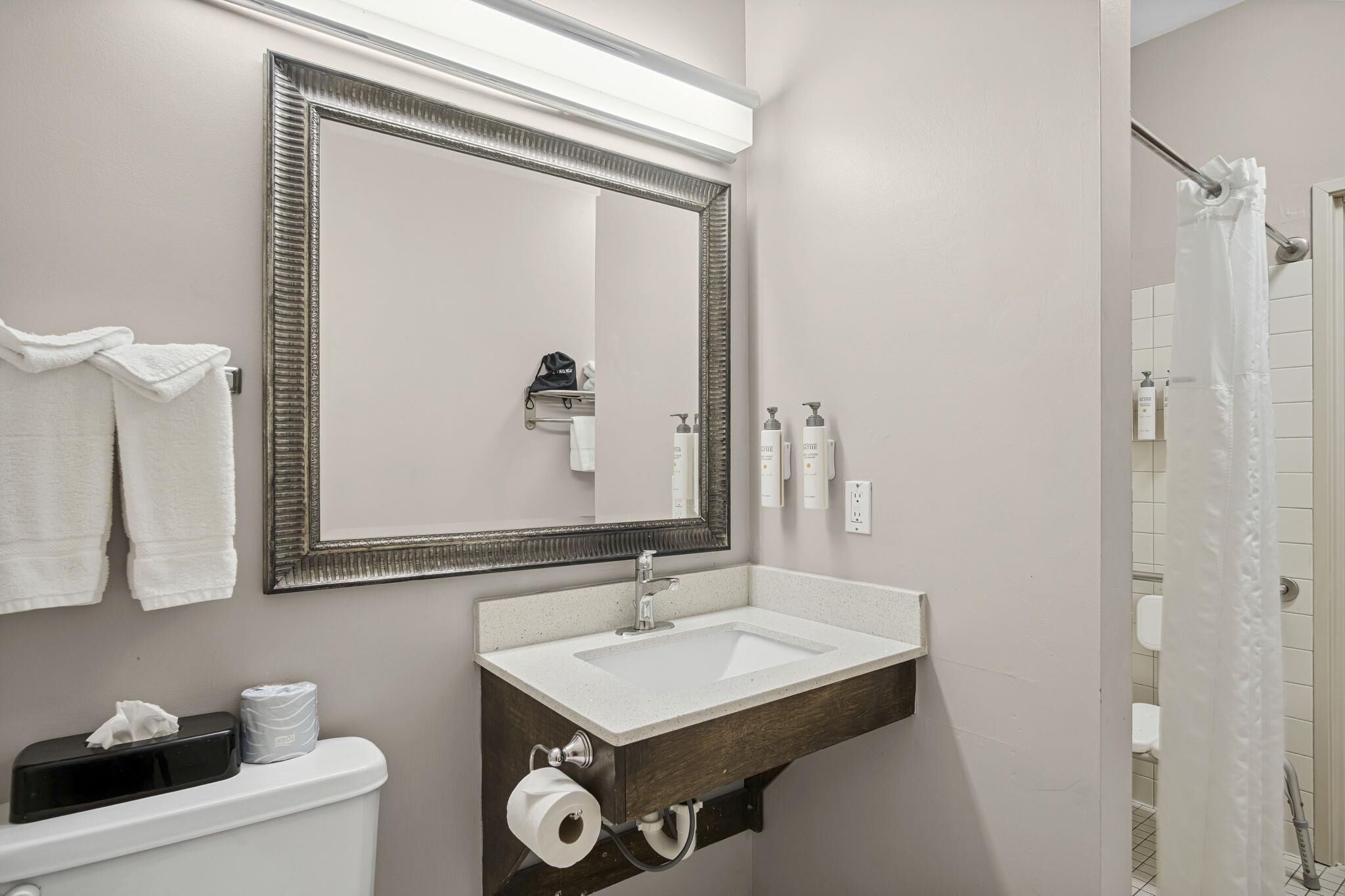 A bathroom with a large mirror, a sink, and a toilet. There are towels hanging on the wall, a shower curtain, and toiletries on the sink.