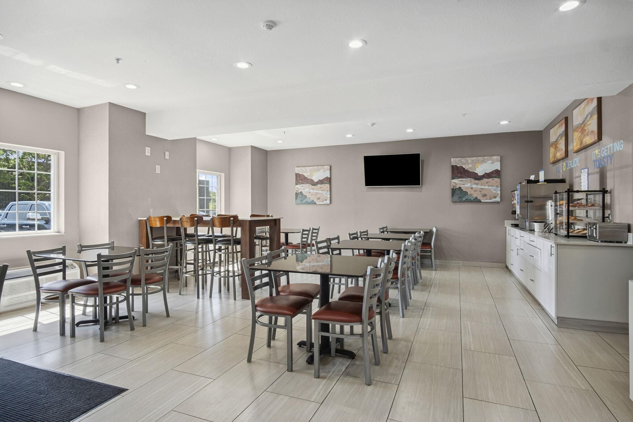 A spacious dining area with multiple tables and chairs, a kitchen counter, and artwork on the walls.