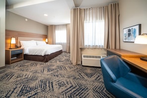 A hotel room with a large bed, a desk, a chair, a nightstand, and a wall-mounted air conditioner.