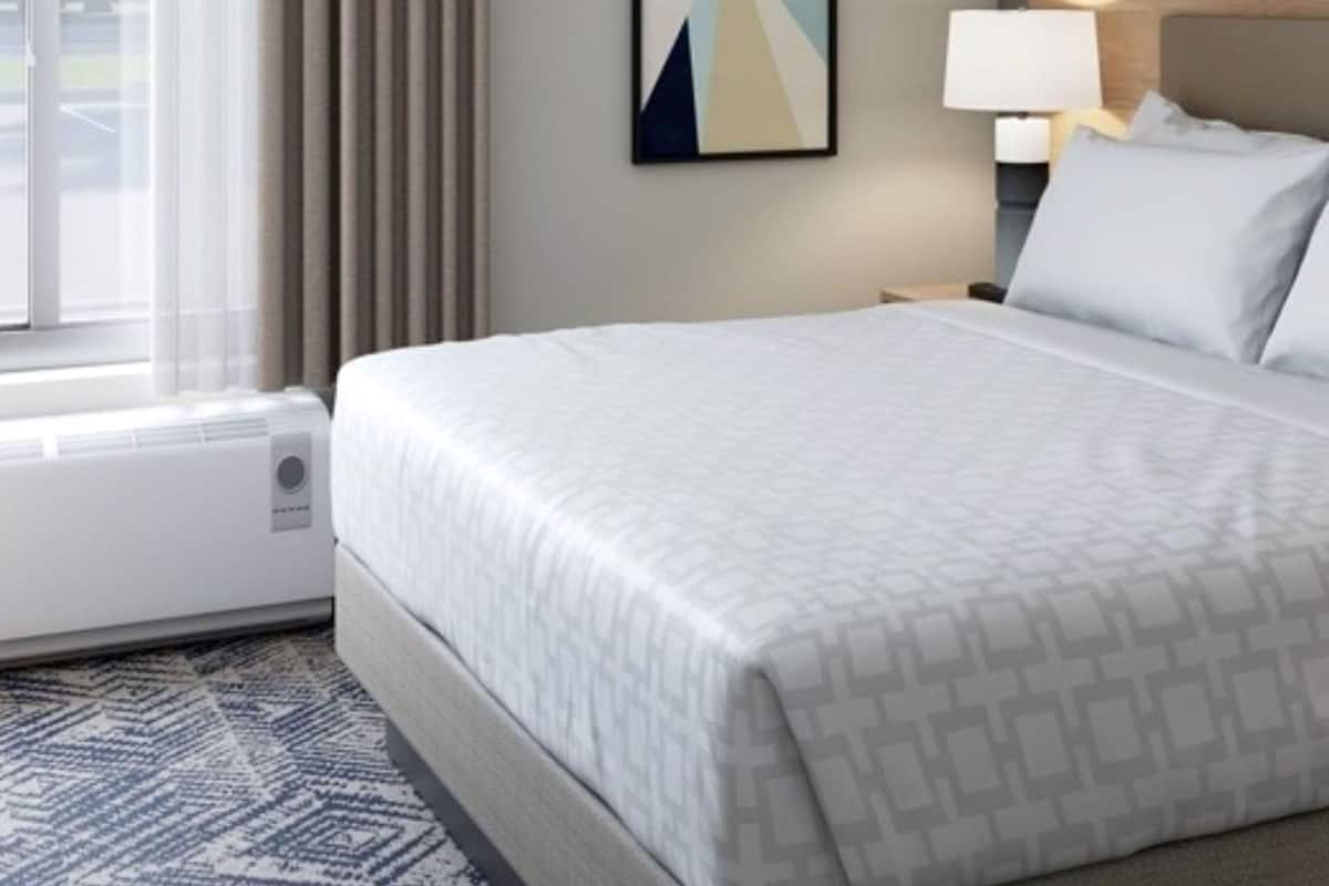A hotel room with a bed, a nightstand, a lamp, a window with curtains, and a wall-mounted artwork.