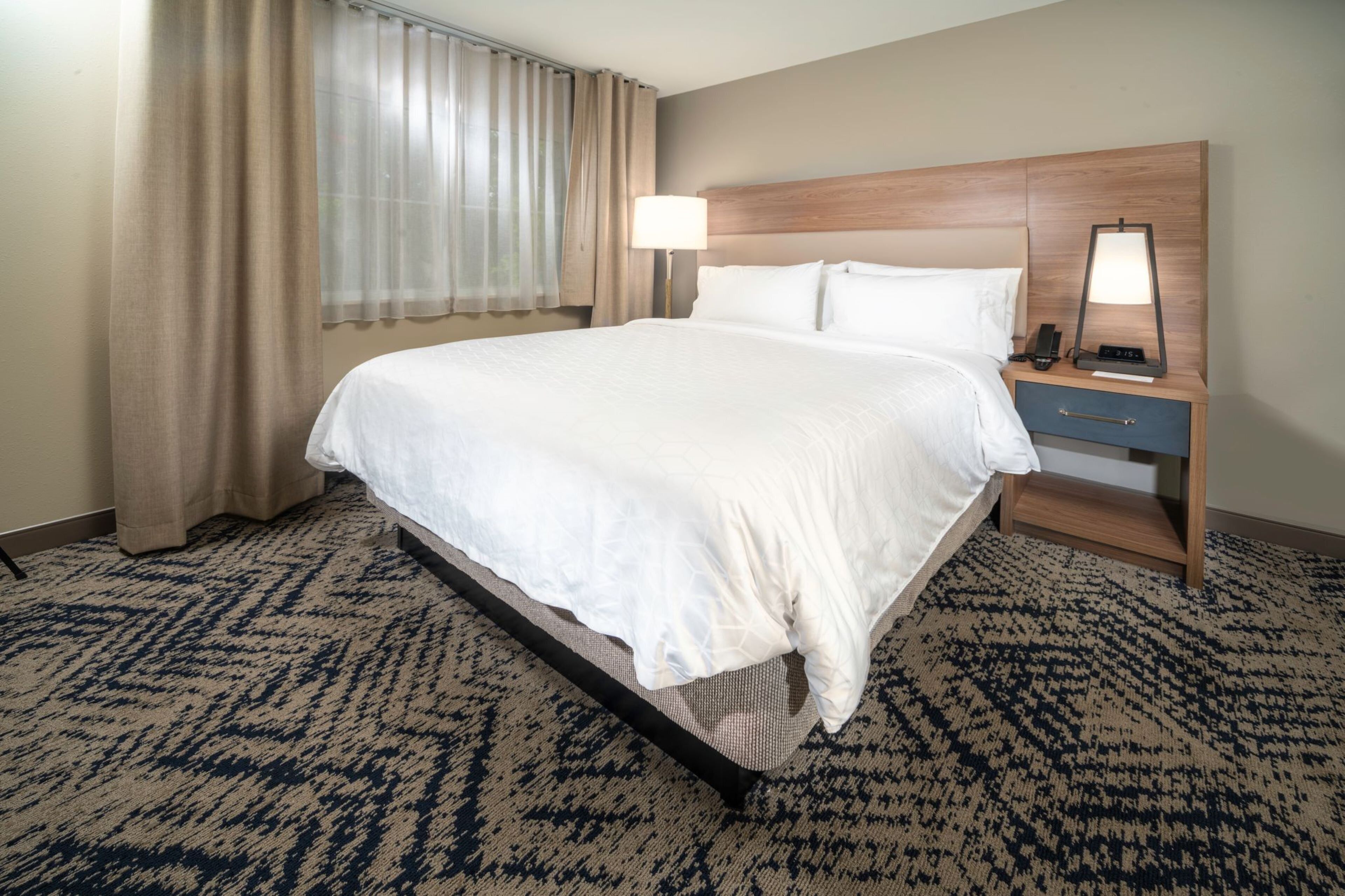 A hotel room with a bed, a nightstand with a lamp, a window with curtains, and a patterned carpet.
