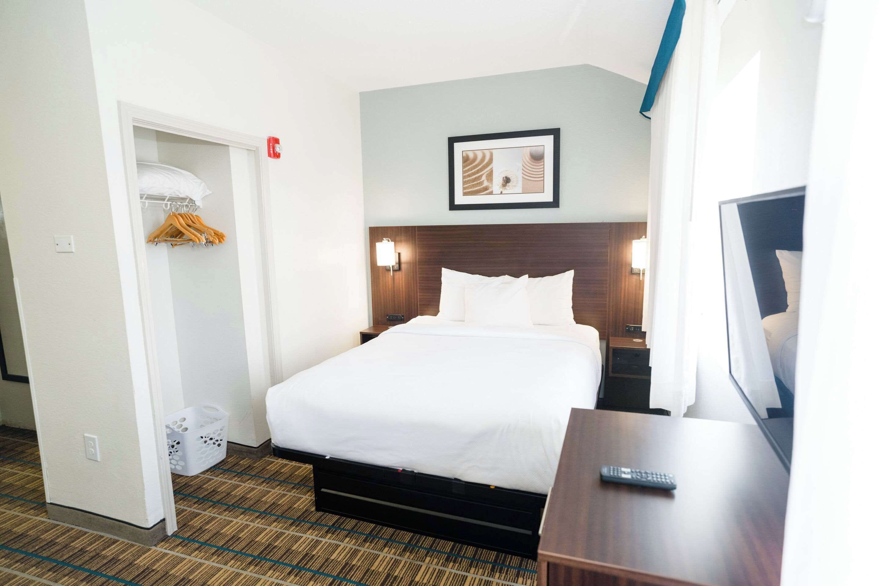 A hotel room with a bed, a wardrobe, a wall-mounted picture, a bedside table, and a remote control.