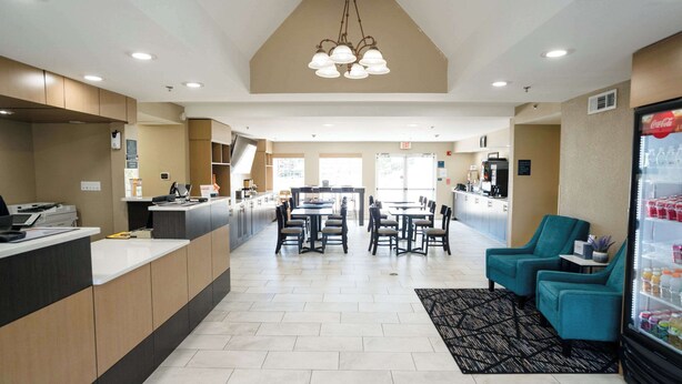 Un hall de hotel con mostrador de recepción, un área de comedor con mesas y sillas, un refrigerador lleno de bebidas y un ventanal que proporciona luz natural.