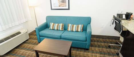 Un sofá color turquesa con almohadones a rayas, una mesa de centro de madera, un cuadro enmarcado en la pared, una lámpara de pie y una kitchenette con microondas y cafetera.