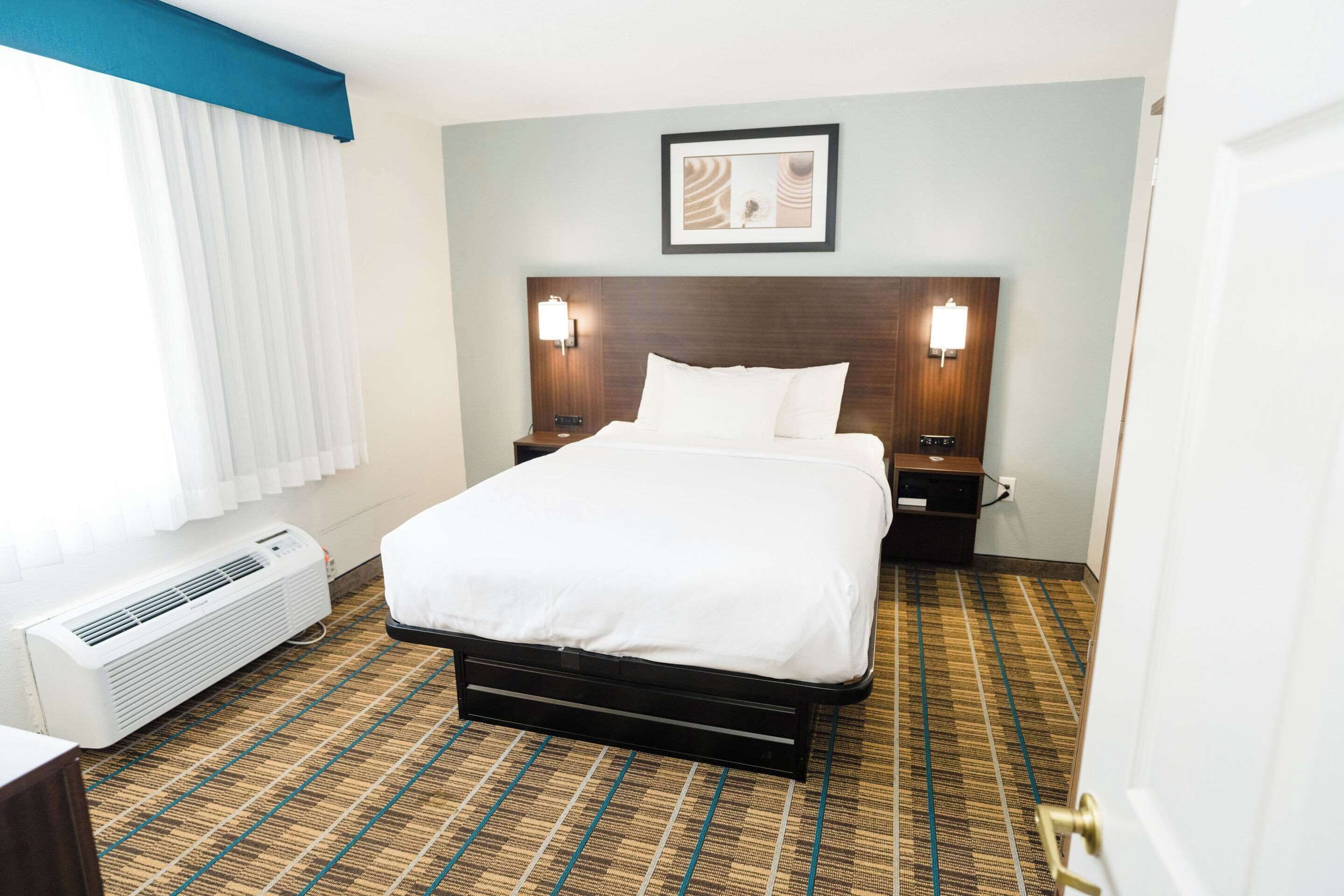 A hotel room with a bed, a wall-mounted light, a heater, and a patterned carpet.
